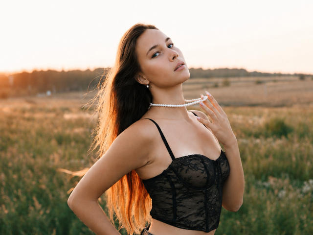 AmmaLoyXX with long brown hair, wearing a black lace bra, holding a pearl necklace, standing in a field at sunset, looking seductive with a slight smile.