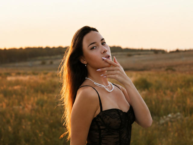 AmmaLoyXX with long brown hair, wearing a black lace top and pearl necklace, touching her lips with her finger, sunlight in her hair, standing in a grassy field.