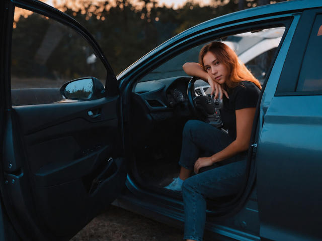 AmmaLoyXX, con cabello naranja, camiseta negra y jeans azules, está sentada en un coche azul con la puerta abierta, apoyando la cabeza en el volante y mirando a la cámara. No se ve la gata..