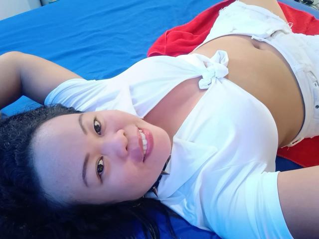 Filipina69-hot with dark curly hair, smiling, lying on blue bed, wearing tied white shirt, exposing small white panties, red blanket, and black choker. Her tits are partially visible.
