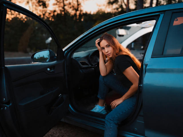 AmmaLoyXX con cabello naranja largo, sentada en un coche azul, con la mano en la cabeza, blusa negra, jeans, zapatillas blancas, puerta abierta, atardecer, árboles en el fondo, expresión seria, sexy, atractiva..