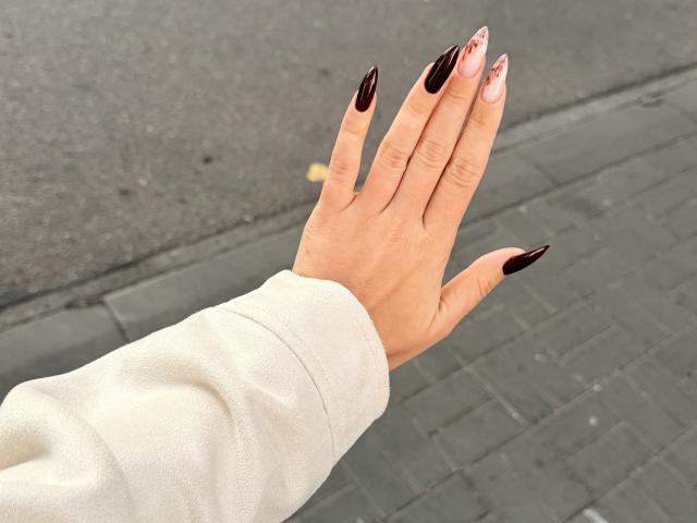 Les ongles longs, pointus, noirs et roses dégradés de SugarrX, mettant en valeur les pointes brillantes, portant une manche blanche, sur fond de trottoir gris et d'asphalte..