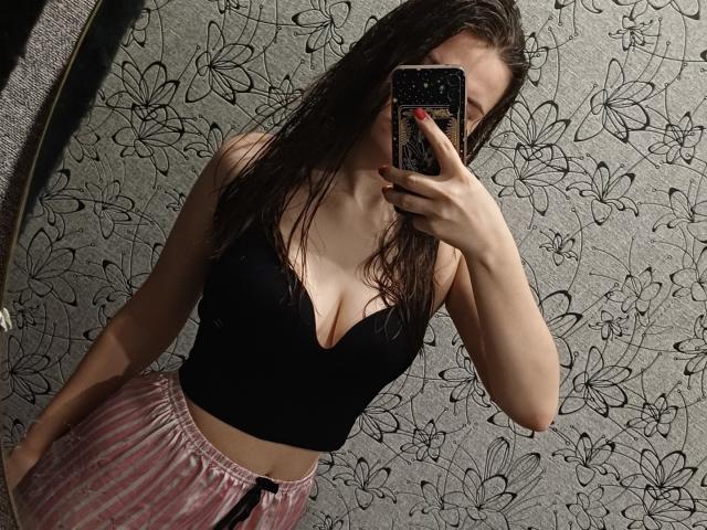 CathrynJacquet with wet brown hair, black bustier, showing her tits, pink velvet pants, holding phone with black floral case, red nails, mirror selfie, standing in dim room.