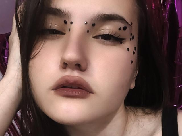 CathrynJacquet has black dots on her eyebrows and nose, with her hand in her brown hair, wearing black eyeliner, and showing her pouty, glossy lips.