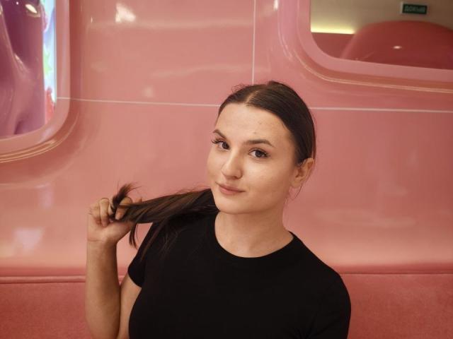 CathrynJacquet with brown hair in a ponytail, holding it with her right hand, wearing a black shirt, smiling slightly, pink glossy background.