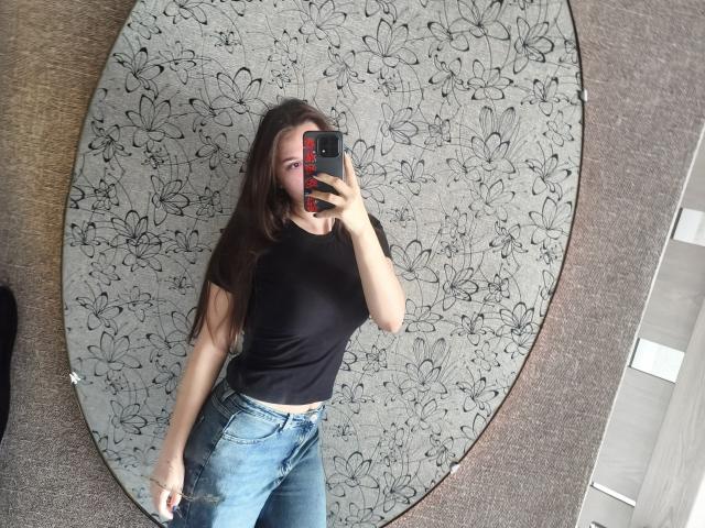 CathrynJacquet with long brown hair, black t-shirt, and blue jeans, standing, taking a mirror selfie, showing her tits and ass, holding a red and black phone.