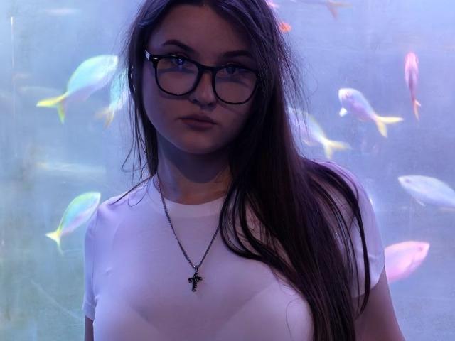 CathrynJacquet with long brown hair, wearing glasses and a white t-shirt, showing her tits, and a cross necklace, standing in front of a blue-tinted aquarium with colorful fish.