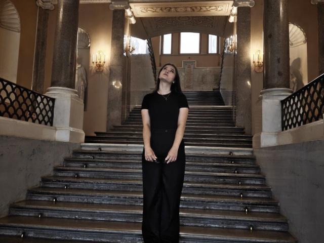 CathrynJacquet stands on marble stairs, wearing a black dress and short sleeves, head tilted up, eyes closed, hands resting on her belly, looking serene and slightly tired.