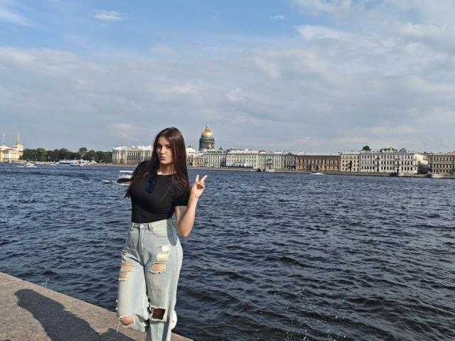 CathrynJacquet with long brown hair, black V-neck, ripped light blue jeans, standing by water, making a peace sign, water and building with dome in background.