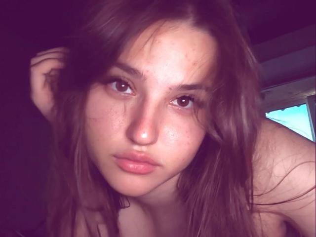 CathrynJacquet with messy brown hair, slightly flushed skin, and serious brown eyes, leaning forward, showing bare shoulders, looking directly at the camera. Her lips are slightly parted.
