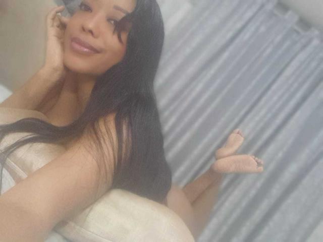 ShantyMarlovy with long black hair, lying on a couch, smiling, holding a phone to her ear, legs up, bare feet, wearing a strapless top, showing her boobs.