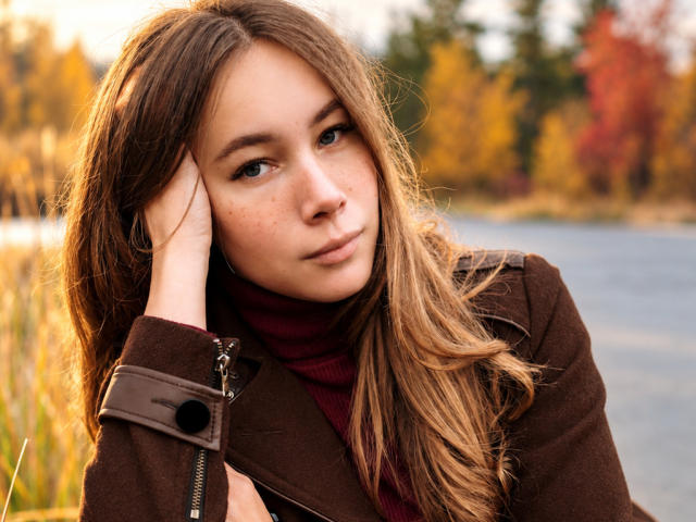 AmmaLoyXX, con cabello castaño largo, pecas y ojos marrones, vestida con una chaqueta marrón y un cuello alto rojo, con la mano en la cabeza, mirando pensativa junto a la orilla del agua. Colores otoñales en el fondo..