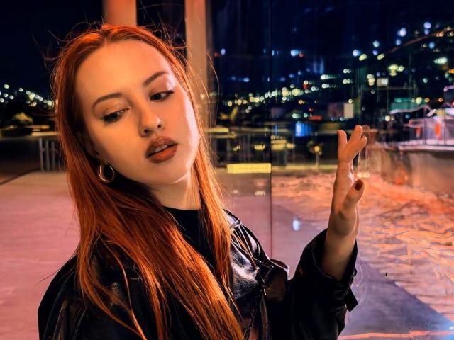 Janna Corsaro mit roten Haaren, die eine schwarze Lederjacke trägt, eine Zigarette hält und nach unten schaut, mit Stadtlichtern im Hintergrund, kleinen Reifenohrringen, leicht geöffneten Lippen, bei Nacht..