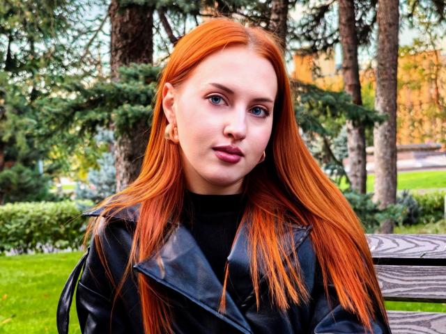 JannaCorsaro with long red hair, wearing a black leather jacket, sitting on a wooden bench, looking at the camera with blue eyes and pink lipstick.