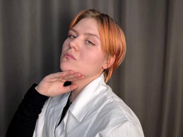 BlytheErby with orange-dyed hair, white shirt, black long-sleeve, hand near chin, slightly pouting, grey curtain background, looking off-camera.