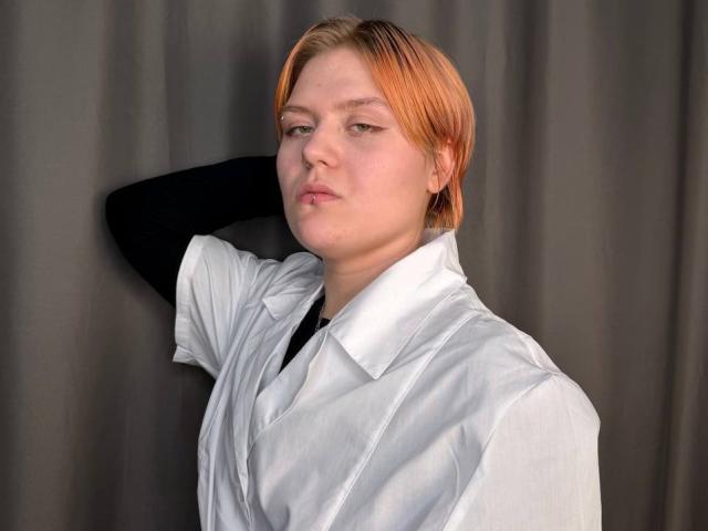 BlytheErby with short orange hair, wearing a white shirt and black sleeve, arms behind head, neutral expression, fair skin, soft lighting, subtle makeup, slight pink lips.