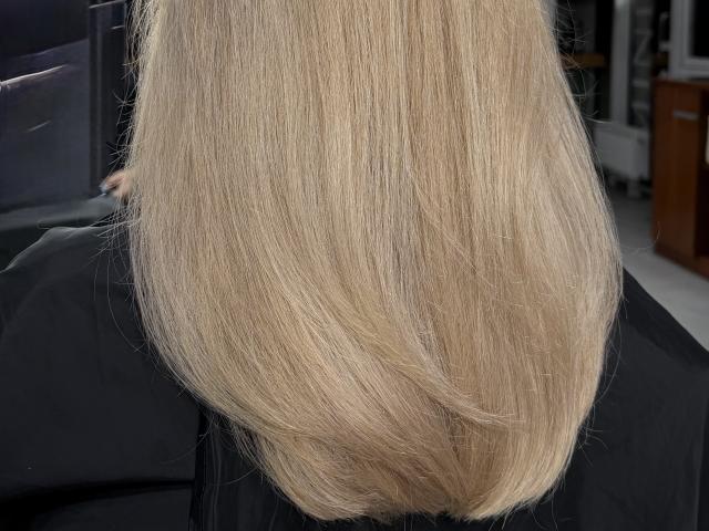 SugarrX's blonde, straight, shoulder-length hair, smooth and shiny, with subtle highlights, covering her black top. Background is blurred and indistinct.