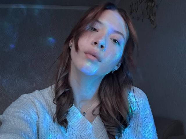 CamillaBoisson with blue-lit face, wavy brown hair, white knit sweater, slight pout, and earrings, looking off-camera. Her tits are subtle under the sweater.