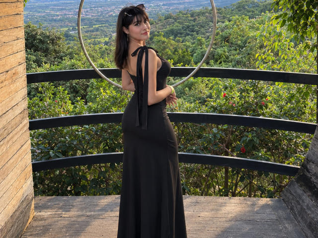AshlyLee se tient dos à la caméra, vêtue d'une robe noire à encolure dégagée, mettant en valeur ses fesses, dans un espace extérieur en bois avec un anneau métallique derrière elle..