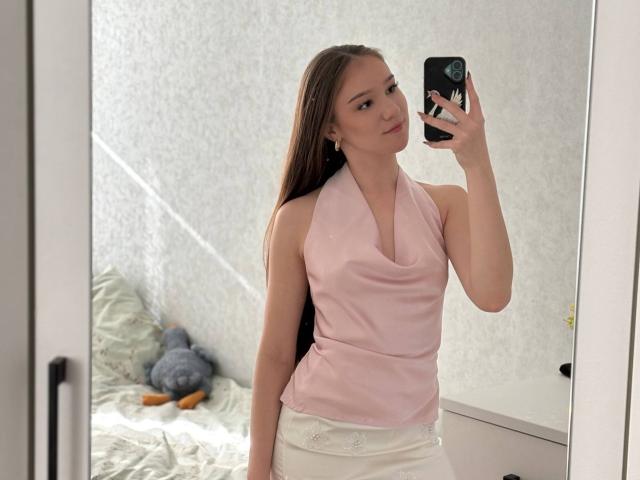 DomingaSierra mit langen braunen Haaren, einem pinken Halstopp, weißen Hosen, die ein Handy hält, ein Selfie vor einem Spiegel, kleine Brüste, schlank, süß, weiße Wand, Bett mit Stofftier im Hintergrund..