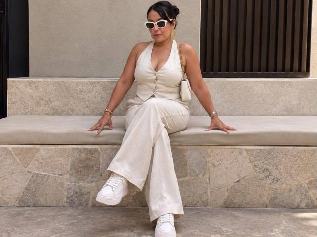 TamaraFoxy, con un sedere importante, indossa un top bianco a fascia, pantaloni bianchi, scarpe da ginnastica bianche e occhiali da sole bianchi, seduta su una panchina di pietra, con le mani sulle ginocchia, capelli neri raccolti in uno chignon..