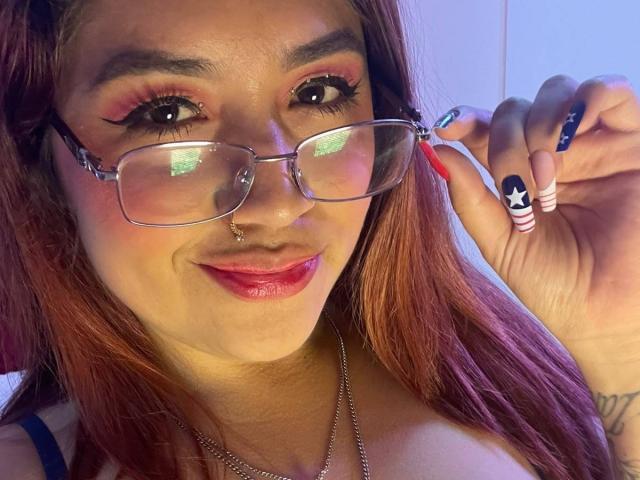 ValmontAlessia, met rood haar, een bril, een neusring, roze lippenstift en nagels met een sterrenpatroon, houdt een bril vast en laat met een schattende glimlach haar borsten zien..