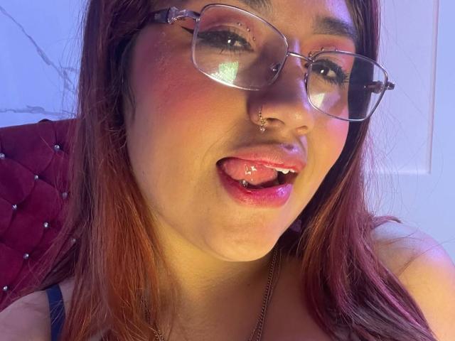 ValmontAlessia with glasses, nose piercings, and tongue piercing, tongue out, red lipstick, and pink blush, showing a bit of saliva, with long brown hair and a red headrest.