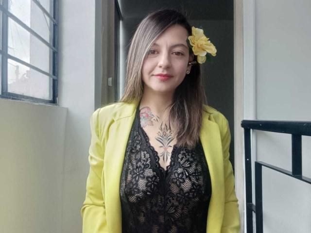 ArianaBlonde with yellow blazer, black lace top, and yellow flower in her brown hair, showing tattoo on chest, standing near window, smiling. Boobs visible through lace.