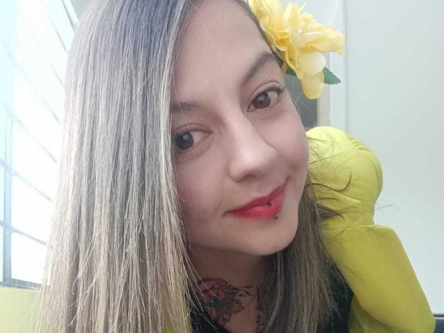 ArianaBlonde has straight blonde hair, a yellow flower in her hair, red lipstick, a lip piercing, and a tattoo on her neck. She's smiling with a yellow pillow behind.