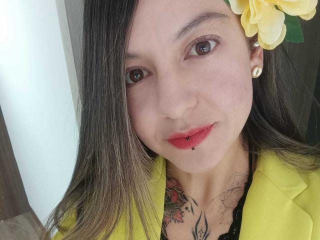 ArianaBlonde with long brown hair, yellow flower, red lipstick, small beauty mark on lower lip, yellow blazer, earrings, tattooed chest, and brown eyes, smiling softly.