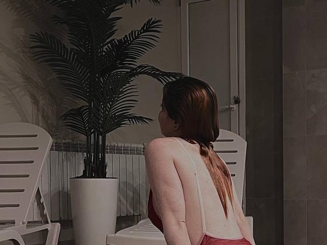LilliamChevalier with long brown hair, back to camera, wearing a red dress with a low back, sitting near a white chair and potted plant in a dim room.