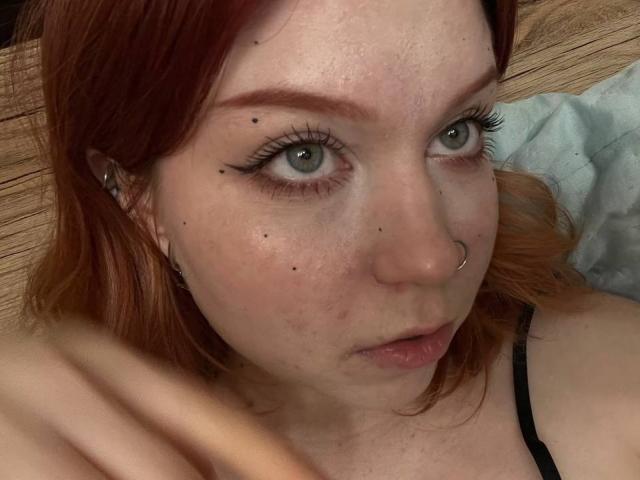 LilliamChevalier has red hair, green eyes, black eyeliner, a nose ring, and several small black moles on her face. Her lips are slightly parted. She's wearing a black strap.