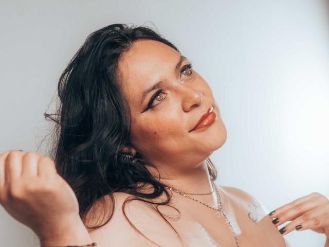 BiancaHottiest met golvend zwart haar, bruine ogen en rode lippenstift, met vuisten omhoog, gekleed in een witte kanten top en een parelketting, met een lichte glimlach..