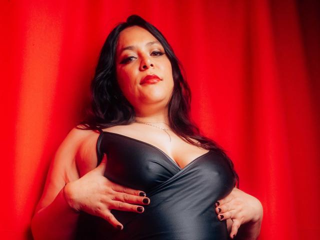 BiancaHottiest com seios grandes, vestindo um vestido de couro preto, segurando os seios, unhas pretas, cortina vermelha, expressão confiante, cabelo preto longo, batom vermelho..