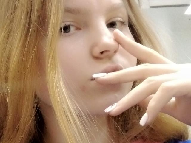 MaximaLasley with light brown hair, pale skin, and white nails, touching her slightly puffy nose with her finger. Her eyes are looking forward. Her lips are slightly parted.