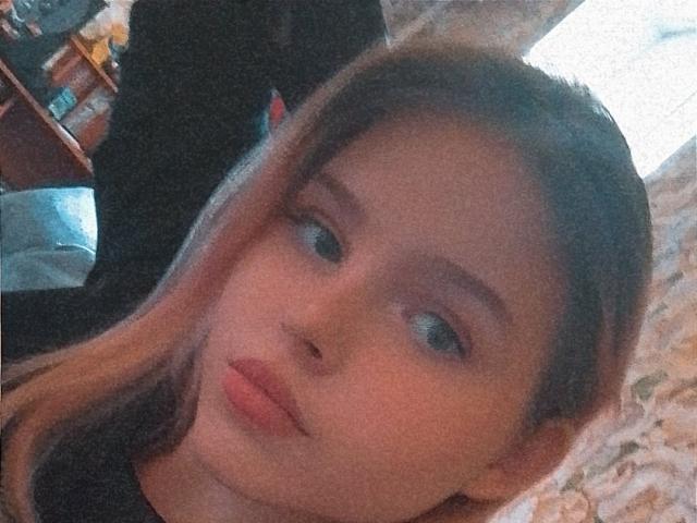 MaximaLasley has light brown hair, blue eyes, and pink lips, looking slightly to the side with a subtle pout, wearing a black top, in a softly lit room.