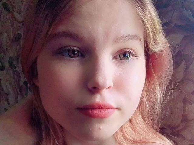 MaximaLasley's face, pale skin, blue eyes, pink lips, blonde hair, slight pout, small zit on her chin, subtle makeup, soft natural light, slight blush, relaxed expression.