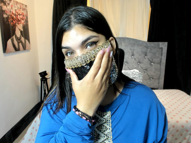 Aniisa Ali mit dunklem Haar, blauem Oberteil, schwarzer Spitzenmaske, Hand, die den Mund bedeckt, blauen Ohrringen, rosafarbenen Nägeln, Armband, sitzend auf einem Bett, grauer Kopfteil, weißer Vorhang, schwarze Wanddekoration..
