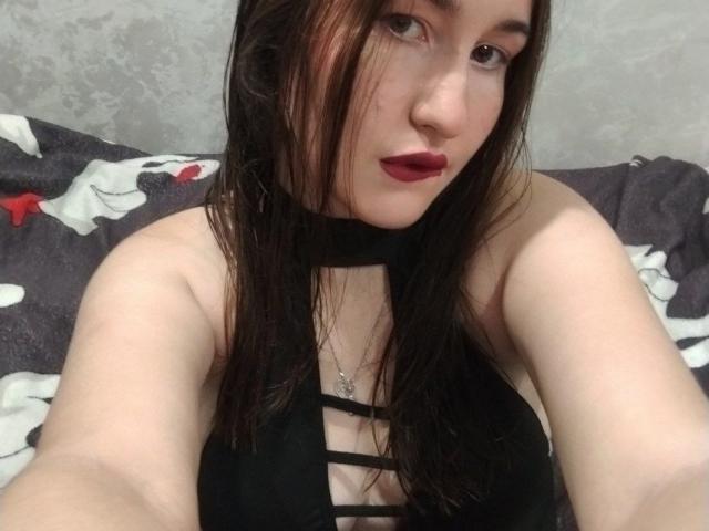 RosalindCutsinger con el cabello castaño mojado, lápiz labial rojo, top negro con tiras, mostrando sus senos, sosteniendo un selfie, piel pálida y un pequeño collar con una cruz, mirando a la cámara..