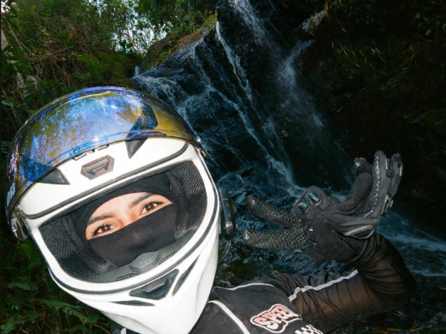KimmyK wearing a white and blue helmet, black face mask, and gloves, with brown eyes visible, standing by a waterfall, making a peace sign with her right hand.