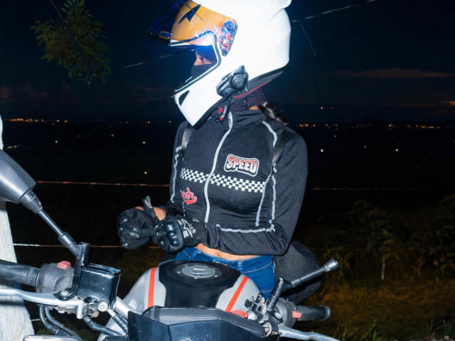 KimmyK with a white helmet, black checkered jacket, and gloves, sitting on a motorcycle at night, wearing blue pants, hands on the handlebars, yellow helmet visor.