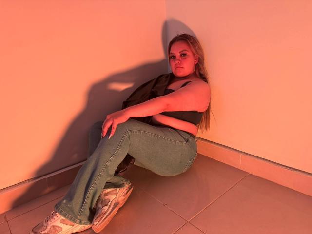 MiaHaruki, com cabelos longos e loiros, blusa de tanque preta, jeans azuis e tênis brancos, sentada encostada em uma parede de canto, pernas cruzadas, braços envolvendo os joelhos, sob iluminação laranja..