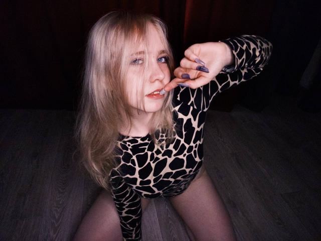 TiffaniMacandog with blonde hair, blue eyes, wearing a black and beige leopard print bodysuit, kneeling on wooden floor, touching her nose with purple-painted fingers, slightly parted lips.