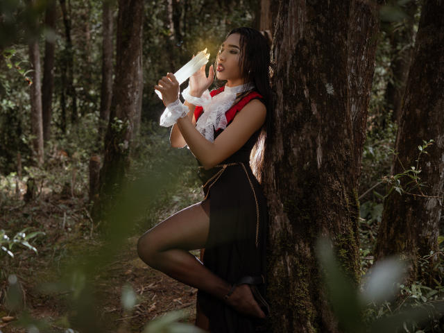 GabySantos leaning against a tree in a forest, holding a glowing wand, wearing a red and black dress with white lace, her black panties visible, dark hair, seductive pose.