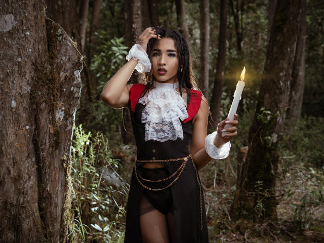 Gaby Santos mit dunklem Haar, einem roten und schwarzen Spitzenkleid, weißen Manschetten, die eine brennende Kerze hält, steht in einem Wald, hat eine Hand auf dem Kopf, wirkt ernst und zeigt ihr Gesäß..