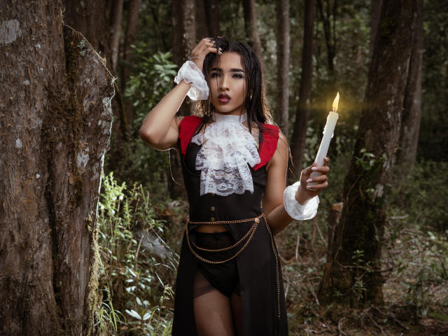GabySantos with dark wet hair, wearing a red and black lace dress, holding a lit candle, standing in a forest, right hand touching her head, looking intense.