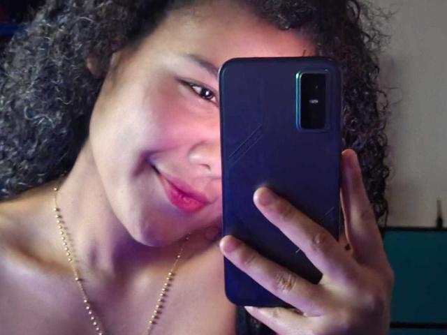 AriaVega with curly black hair, holding a blue phone, smiling with pink lipstick, wearing a gold necklace, showing her ass with her shoulder.