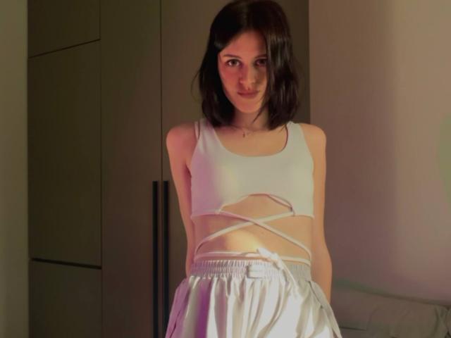 LachelleCaraker with short brown hair, wearing a white crop top with cutouts and white skirt, standing in a room, looking at the camera with a slight smile.