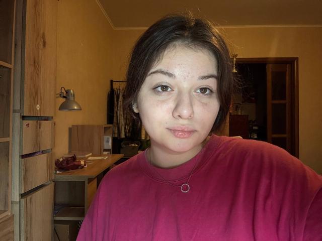 KendalBeulah, uma garota rechonchuda com acne, olhos castanhos e cabelo escuro, vestindo uma camisa vermelha, fazendo beicinho com os lábios levemente entreabertos e usando um colar simples, em um quarto com pouca luz..