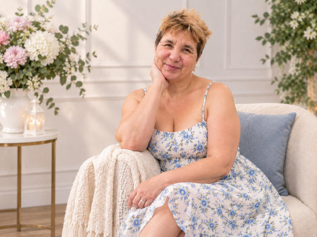 Adelina Luna, eine Frau mittleren Alters mit kurzen, blonden Haaren, trägt ein blaues Kleid mit Blumenmuster und sitzt auf einem weißen Sofa. Sie lächelt, hat eine Hand an der Wange, und es gibt eine weiße Spitzendecke, ein blaues Kissen und eine Blumenvase..